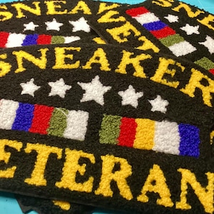 May include: Black doormats with yellow text that reads "SNEAKER VETERAN". Each mat features white stars and colorful stripes in red, white, blue, and green. The mats are made of a textured material.
