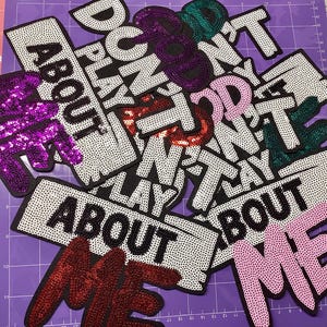 Może przedstawiać: Kolekcja naszywek z cekinami z napisami "DON'T PLAY ABOUT ME" i "DON'T PLAY ABOUT" w różnych kolorach. Naszywki są czarne, białe, fioletowe, czerwone, różowe i zielone, z detalami z cekinów i koralików.