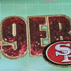 Puede incluir: Un parche grande con lentejuelas con la palabra "49ERS" en lentejuelas rojas con ribete dorado brillante. Un parche ovalado con el logotipo del equipo está debajo. Los parches son probablemente para ropa.
