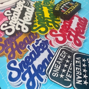 May include: A collection of embroidered patches in various colors, including pink, yellow, green, red, purple, and blue. The patches feature the words "Sneaker Head" in a script font and "Sneaker Veteran".