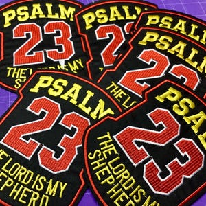 Może przedstawiać: Haftowane czarne naszywki z napisem "PSALM 23" w kolorze żółtym i czerwonym. Na naszywkach wyhaftowano również frazę "THE LORD IS MY SHEPHERD". Naszywki mają kształt tarczy.