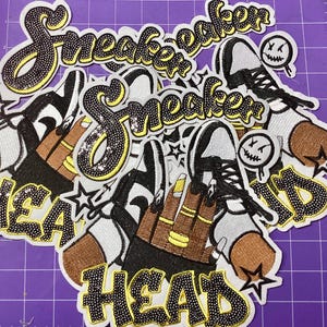 May include: A collection of embroidered patches featuring the text Sneaker Head in black with yellow outlines. The design includes illustrations of high-top trainers, a hand with yellow rings, and small circular faces with stitched mouths.