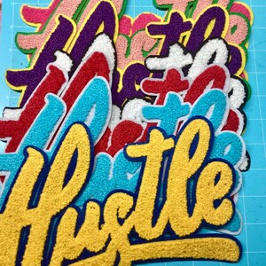 以下が含まれることがあります： 黄色、ターコイズ、赤、白、紫、ピンクなど、さまざまな色の「Hustle」という文字がデザインされた、重ねられたシェニール織のワッペン。各ワッペンは厚みのある質感の表面で、太い筆記体フォントと縁取りが特徴です。