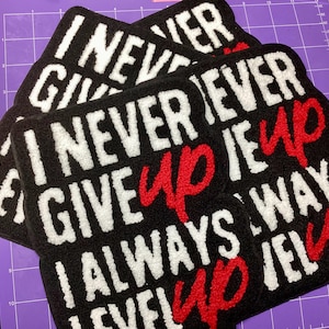 May include: Black rectangular rugs with the text "I NEVER GIVE UP" in white and red. The rugs are made of a soft, textured material and are arranged in a stack.