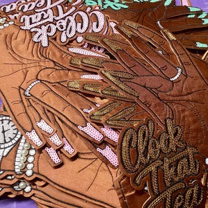 May include: Brown fabric patches in the shape of hands with long, gold-colored nails. The patches have black outlines and are embellished with sequins and pearls. Text on the patches reads "Clock That Tea".