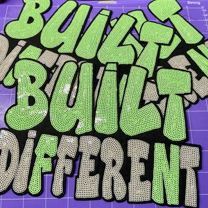 May include: Black patches with the words "BUILT DIFFERENT" in a bold, bubble-style font. The letters are outlined in black, with the word "BUILT" in light green and "DIFFERENT" in gray. The letters are filled with small dots.