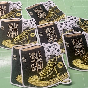 May include: A collection of embroidered patches featuring a black high-top sneaker and a book. The book has the words "WALK WITH G+D" in gold lettering. The sneaker is black with gold accents and a star logo. The patches are on a green surface.