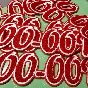 Op de afbeelding: Een verzameling rode viltletters en -cijfers met witte randen, die "OO OOP" en andere combinaties spellen. De items liggen op een groene ondergrond, waarschijnlijk voor knutsel- of personalisatieprojecten.