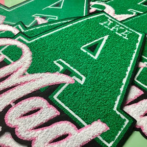 May include: Close-up of several green and white embroidered patches. The patches feature the Greek letters "AKA" and a large letter "A" with a black triangle. The letters are outlined in white and pink.