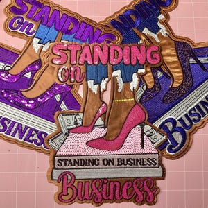 Pode incluir: Patches bordados com o texto "STANDING ON BUSINESS". Os patches apresentam sapatos de salto alto, pilhas de dinheiro e as palavras "STANDING ON" e "BUSINESS" em várias cores, incluindo rosa, roxo e castanho.