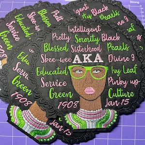 以下が含まれることがあります： 緑色のメガネをかけた黒人女性が描かれた刺繍パッチ。 「AKA」、「Educated」、「Service」などの文字が周囲に書かれています。 パッチは黒色で、緑、ピンク、白のアクセントがあります。 年号の「1908」も表示されています。