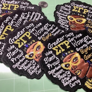 May include: Black embroidered patches with the text "Greater Unity Service Women" and "Sigma Gamma Rho 1922". The patches feature a woman's face with gold-coloured glasses and text like "Woke", "Black", and "Proud".