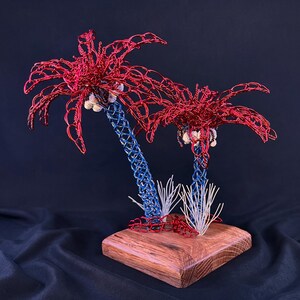 May include: A decorative sculpture featuring two palm trees crafted from red and blue wire. The palm fronds are made of red wire, the trunks are blue wire, and small white shells are attached beneath the fronds. The trees sit on a wooden base.