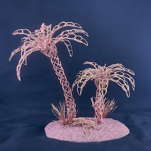 May include: Two palm tree sculptures crafted from thin copper wire, featuring intricate lattice-patterned trunks and arched fronds. The trees are mounted on a circular, textured base with small wire tufts at the base of each trunk.