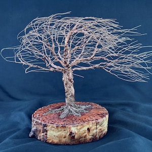 May include: A sculpture of a tree crafted from thin, metallic copper-colored wire, featuring a twisted trunk and branches that extend horizontally to one side. The wire tree is mounted on a circular, light-brown wooden base with visible bark.