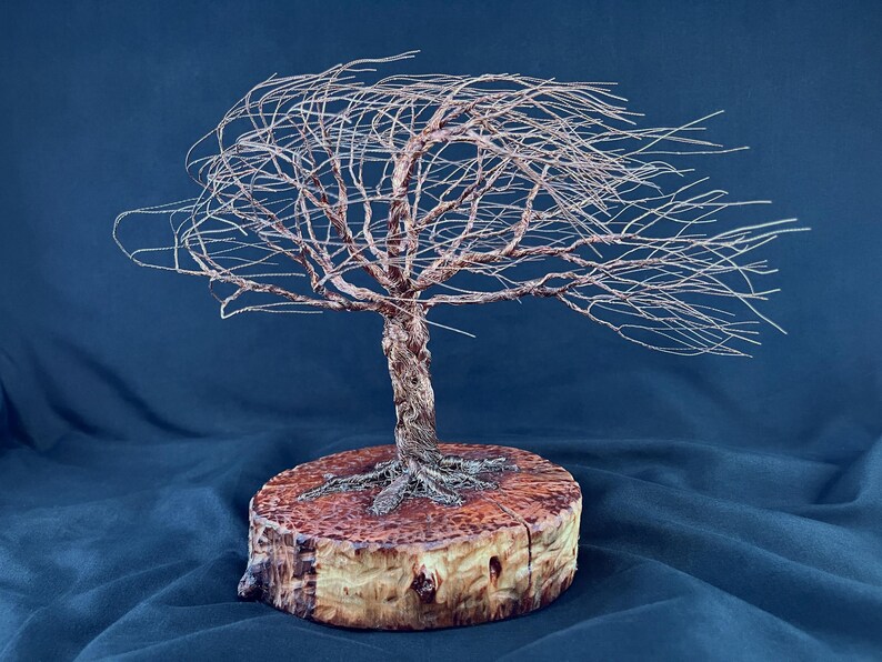 May include: A sculpture of a tree crafted from thin, metallic copper-colored wire, featuring a twisted trunk and branches that extend horizontally to one side. The wire tree is mounted on a circular, light-brown wooden base with visible bark.