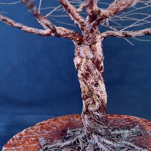 May include: A sculpture of a tree crafted from twisted copper wire, featuring a thick, textured trunk and intricate branches that extend outward. The base of the tree is anchored to a circular, polished wooden disc with a visible grain pattern.