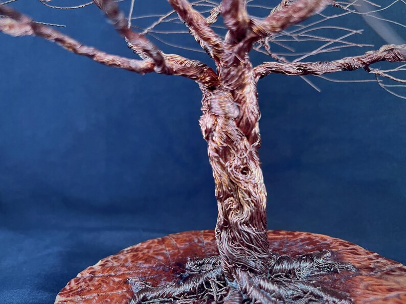 May include: A sculpture of a tree crafted from twisted copper wire, featuring a thick, textured trunk and intricate branches that extend outward. The base of the tree is anchored to a circular, polished wooden disc with a visible grain pattern.