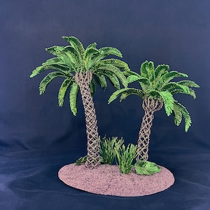 May include: A miniature sculpture featuring two palm trees with textured brown trunks and vibrant green fronds, set on a circular, sandy-colored base. Small patches of green grass are positioned at the base of the trees, creating a tropical island scene.