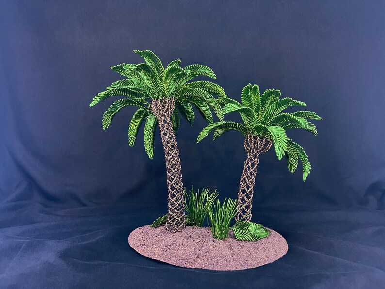 May include: A miniature sculpture featuring two palm trees with textured brown trunks and vibrant green fronds, set on a circular, sandy-colored base. Small patches of green grass are positioned at the base of the trees, creating a tropical island scene.