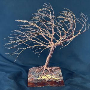 Puede incluir: Una escultura de alambre de un &aacute;rbol azotado por el viento con ramas finas de color cobre que se extienden hacia la izquierda. El tronco est&aacute; formado por alambre trenzado anclado en una base de madera rectangular con corteza visible.