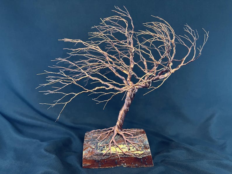 Puede incluir: Una escultura de alambre de un &aacute;rbol azotado por el viento con ramas finas de color cobre que se extienden hacia la izquierda. El tronco est&aacute; formado por alambre trenzado anclado en una base de madera rectangular con corteza visible.