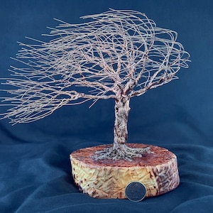 May include: A wire sculpture of a wind-swept tree made from thin copper-colored metal strands. The tree is mounted on a circular wooden base with a visible US quarter coin placed at the front edge for scale, showing the relative size of the sculpture.