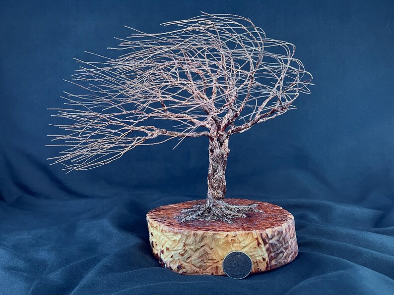 May include: A wire sculpture of a wind-swept tree made from thin copper-colored metal strands. The tree is mounted on a circular wooden base with a visible US quarter coin placed at the front edge for scale, showing the relative size of the sculpture.
