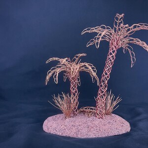 May include: Two palm tree sculptures crafted from thin copper wire, featuring intricate woven trunks and fronds. The trees are mounted on a circular, textured base covered in fine, light-colored sand, with small tufts of copper wire grass at the base.