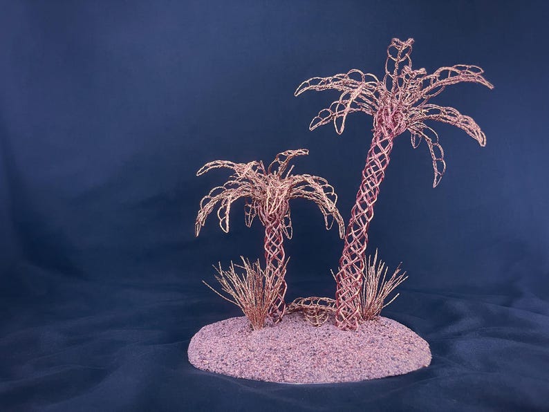 May include: Two palm tree sculptures crafted from thin copper wire, featuring intricate woven trunks and fronds. The trees are mounted on a circular, textured base covered in fine, light-colored sand, with small tufts of copper wire grass at the base.
