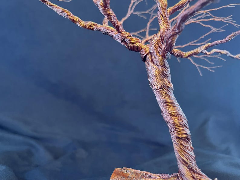 Puede incluir: Una escultura de &aacute;rbol hecha de alambre de cobre trenzado, con un tronco grueso y entrelazado que se ramifica en numerosas ramas finas y delicadas. El metal tiene un acabado c&aacute;lido e iridiscente con tonos bronce, dorados y p&uacute;rpuras.