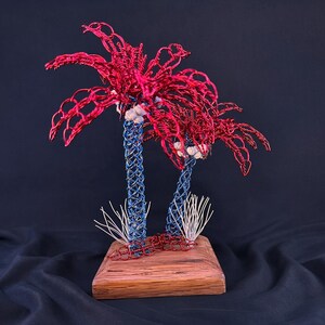 May include: A decorative sculpture featuring two palm trees crafted from twisted wire. The trunks are made of blue wire, while the palm fronds are constructed from red wire. The trees are mounted on a rectangular wooden base with white wire accents.