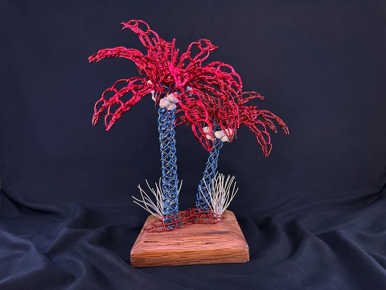 May include: A decorative sculpture featuring two palm trees crafted from twisted wire. The trunks are made of blue wire, while the palm fronds are constructed from red wire. The trees are mounted on a rectangular wooden base with white wire accents.