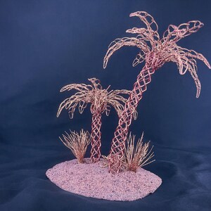 May include: A sculpture featuring two palm trees crafted from copper-colored wire, set on a textured, oval-shaped base. The trees have intricate, woven trunks and delicate, spreading fronds, accompanied by small, spiky wire tufts at the base.