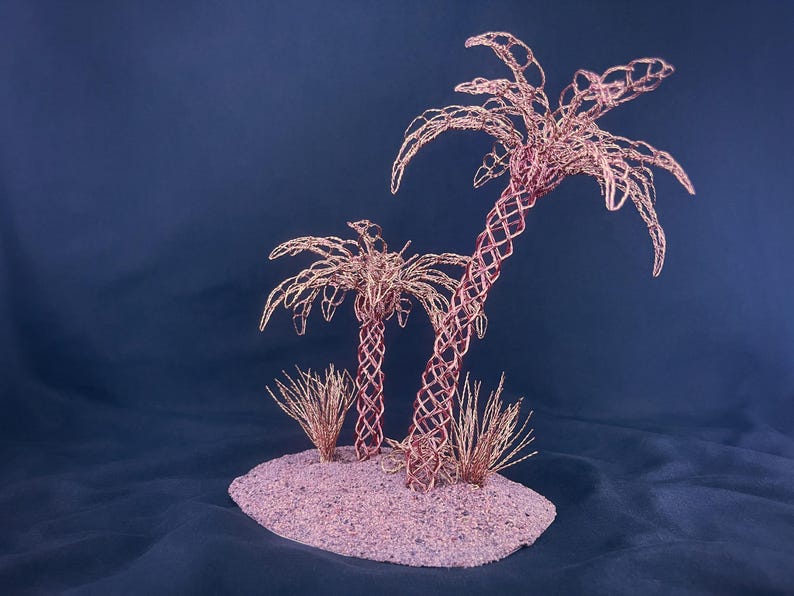 May include: A sculpture featuring two palm trees crafted from copper-colored wire, set on a textured, oval-shaped base. The trees have intricate, woven trunks and delicate, spreading fronds, accompanied by small, spiky wire tufts at the base.