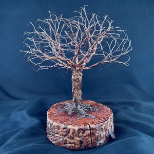 May include: A wire sculpture of a leafless tree with a textured, metallic copper-colored trunk and branches, featuring intricate root details at the base. The sculpture is mounted on a circular, polished wooden log slice with a visible vertical crack.