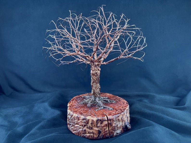 May include: A wire sculpture of a leafless tree with a textured, metallic copper-colored trunk and branches, featuring intricate root details at the base. The sculpture is mounted on a circular, polished wooden log slice with a visible vertical crack.