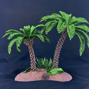 May include: Two wire-sculpted palm trees with vibrant green metallic fronds and textured brown trunks, set on a sandy-colored base. A silver United States quarter coin is placed at the base for scale, showing the relative size of the sculpture.
