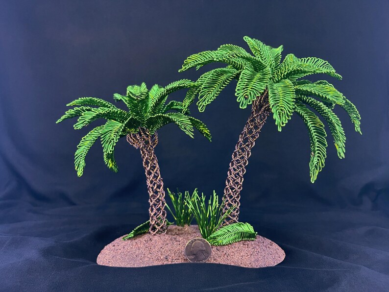 May include: Two wire-sculpted palm trees with vibrant green metallic fronds and textured brown trunks, set on a sandy-colored base. A silver United States quarter coin is placed at the base for scale, showing the relative size of the sculpture.