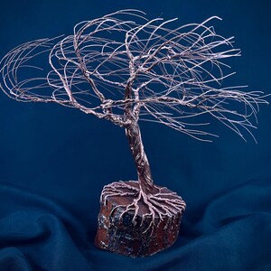 Puede incluir: Una escultura de &aacute;rbol hecha de alambre de cobre fino, con un tronco retorcido y ramas extendidas hacia afuera. Las ra&iacute;ces de alambre est&aacute;n ancladas en una base de madera circular de color marr&oacute;n oscuro con una corteza rugosa.