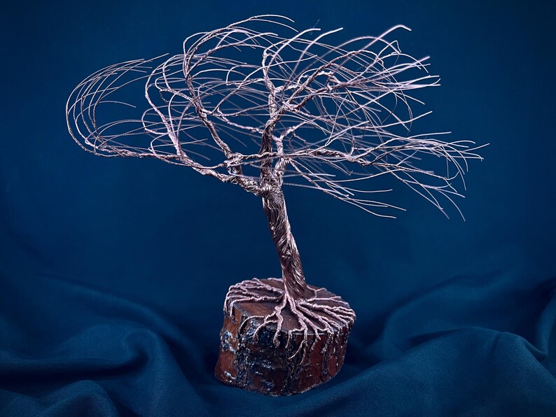 Puede incluir: Una escultura de &aacute;rbol hecha de alambre de cobre fino, con un tronco retorcido y ramas extendidas hacia afuera. Las ra&iacute;ces de alambre est&aacute;n ancladas en una base de madera circular de color marr&oacute;n oscuro con una corteza rugosa.