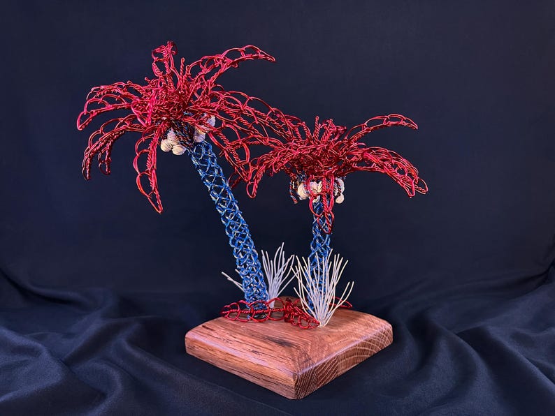 May include: A decorative sculpture featuring two palm trees crafted from red and blue wire. The palm fronds are made of red wire, the trunks are blue wire, and small white shells are attached beneath the fronds. The trees sit on a wooden base.