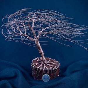 Puede incluir: Una escultura de alambre de cobre de un &aacute;rbol azotado por el viento con ramas finas y fluidas que se extienden hacia un lado. El &aacute;rbol est&aacute; anclado a una base de madera circular con ra&iacute;ces visibles. Una moneda est&aacute; colocada en la base como referencia.