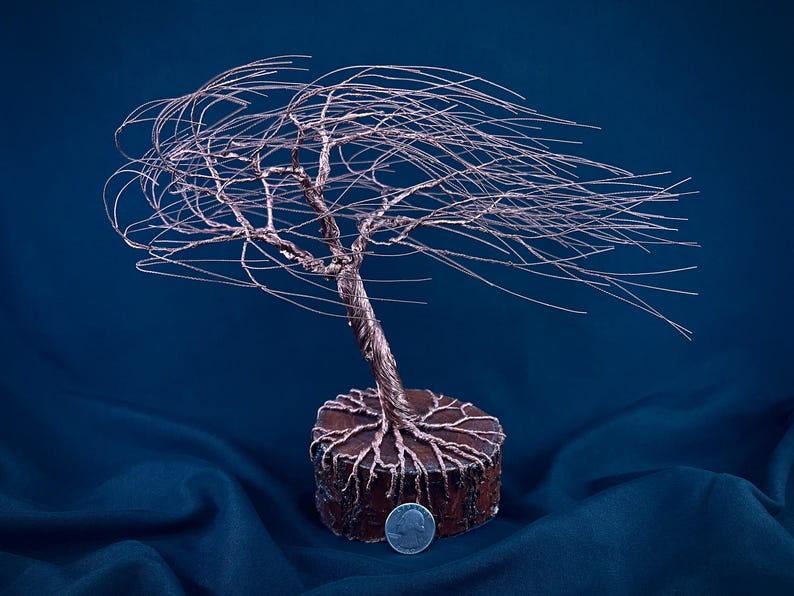Puede incluir: Una escultura de alambre de cobre de un &aacute;rbol azotado por el viento con ramas finas y fluidas que se extienden hacia un lado. El &aacute;rbol est&aacute; anclado a una base de madera circular con ra&iacute;ces visibles. Una moneda est&aacute; colocada en la base como referencia.