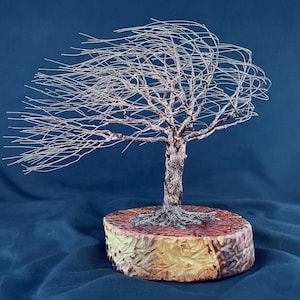 May include: A wire sculpture of a tree with thin, metallic branches extending to one side as if blown by a strong wind. The tree is anchored to a thick, circular wooden base with a textured, light brown bark edge and a smooth, reddish-brown top.