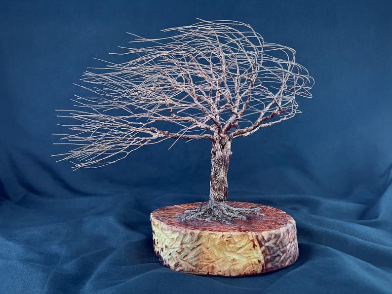 May include: A wire sculpture of a tree with thin, metallic branches extending to one side as if blown by a strong wind. The tree is anchored to a thick, circular wooden base with a textured, light brown bark edge and a smooth, reddish-brown top.