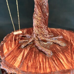May include: A decorative sculpture featuring a tree crafted from twisted copper wire, with a small swing suspended from its branches. The tree is mounted on a circular, polished wooden base that displays a prominent, natural wood grain pattern.