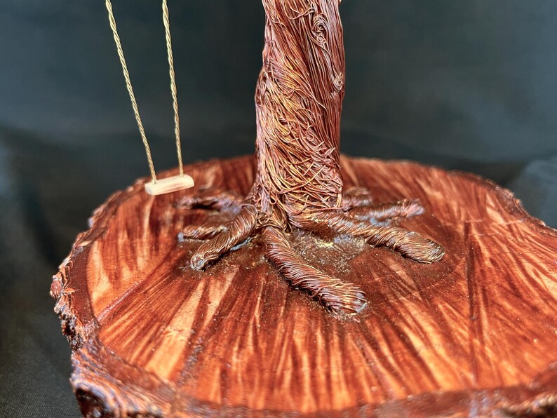 May include: A decorative sculpture featuring a tree crafted from twisted copper wire, with a small swing suspended from its branches. The tree is mounted on a circular, polished wooden base that displays a prominent, natural wood grain pattern.