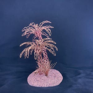 May include: A decorative sculpture featuring two palm trees crafted from thin, metallic copper-colored wire. The trees are mounted on a circular, textured base that is covered in fine, light pink sand, creating a cohesive and detailed miniature display.