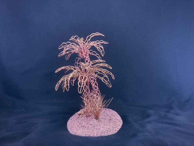 May include: A decorative sculpture featuring two palm trees crafted from thin, metallic copper-colored wire. The trees are mounted on a circular, textured base that is covered in fine, light pink sand, creating a cohesive and detailed miniature display.